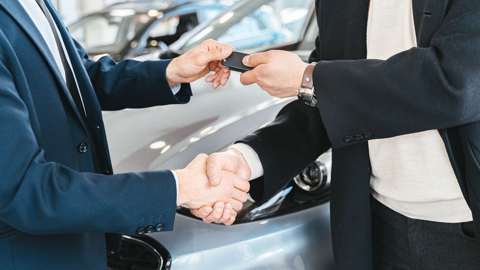 A buyer and an agent shake hands over car keys beside a new car